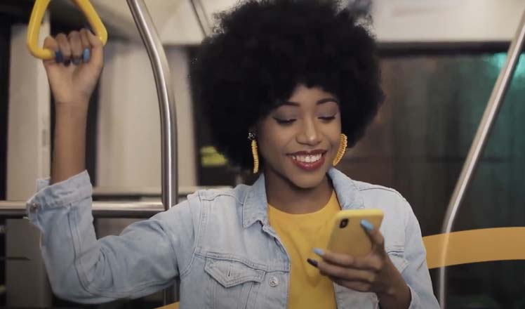 African american young woman using a banking app built with Pismo platform on her mobile phone while taking the tube in a yellow t-shirt